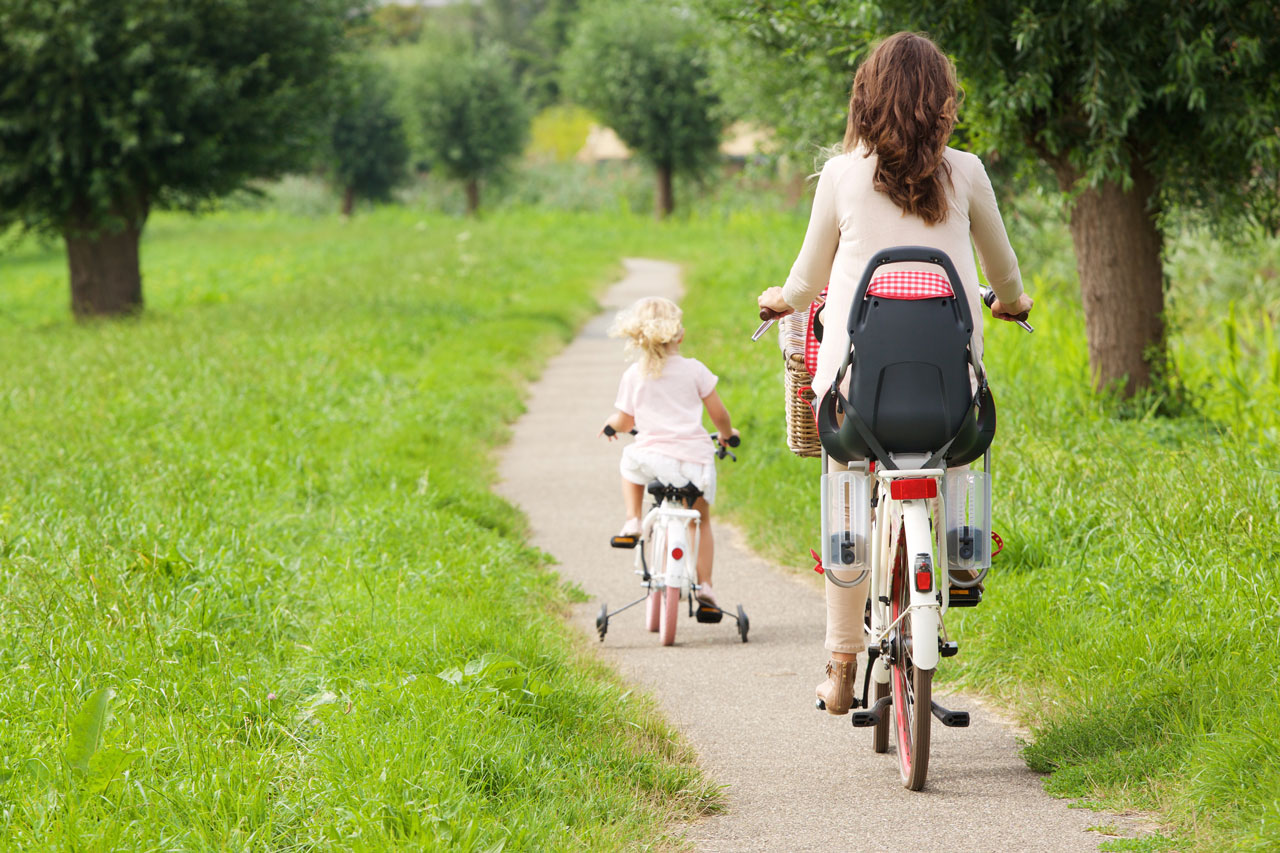 Kind fietsend op fiets met zijwieltjes op smal pad in de natuur. Moeder fietst er achter.